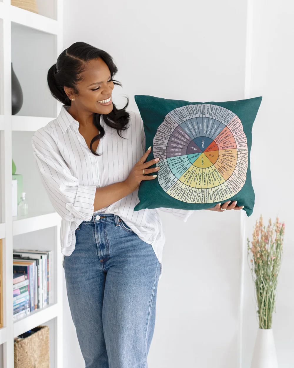 Karina Jackson, standing in front of her bookshelves, laughing as she holds a green cushion printed with an emotion wheel