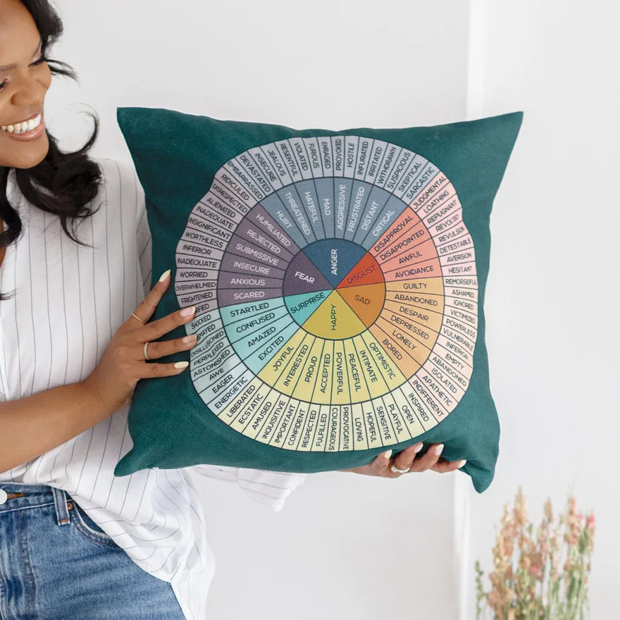 Close-up of Karina Jackson's cushion, printed with an emotion wheel: fear, anger, sadness, joy and their shades; her hands hold it, her smile at the edge of the frame
