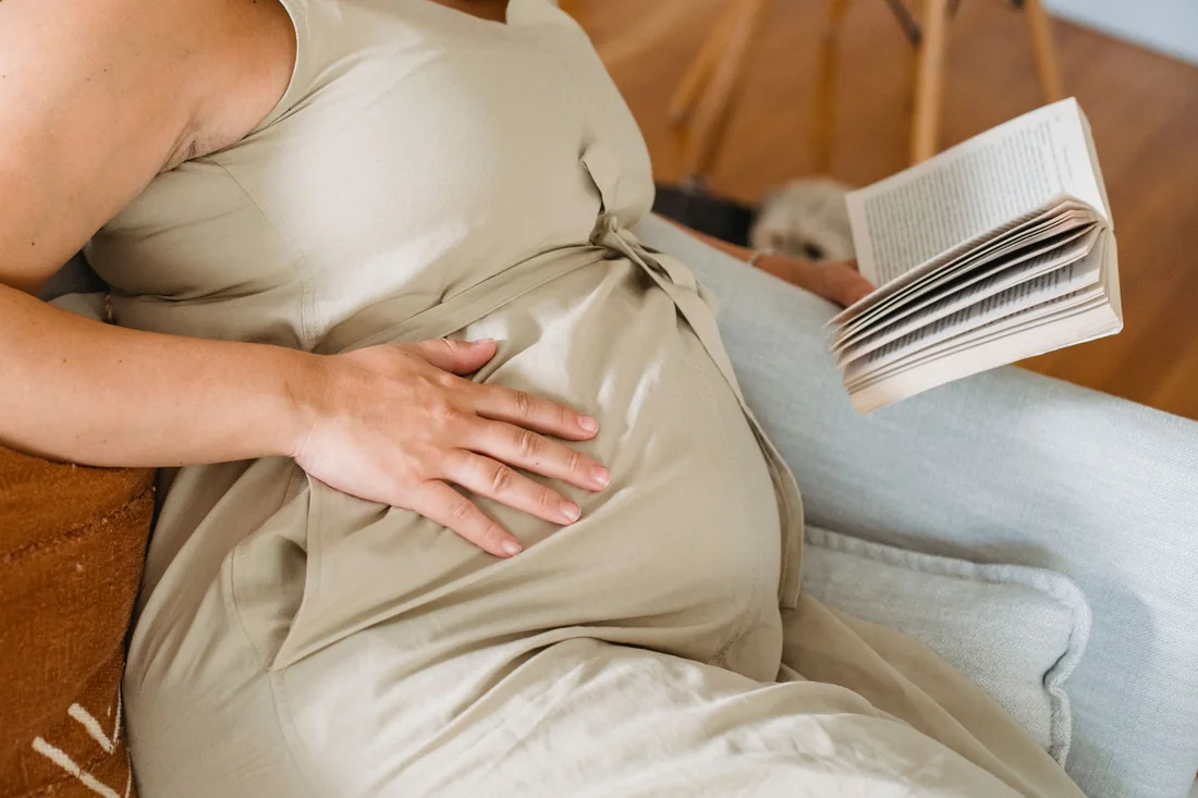 A pregnant woman seated on her sofa, one hand on her belly, an open book in the other