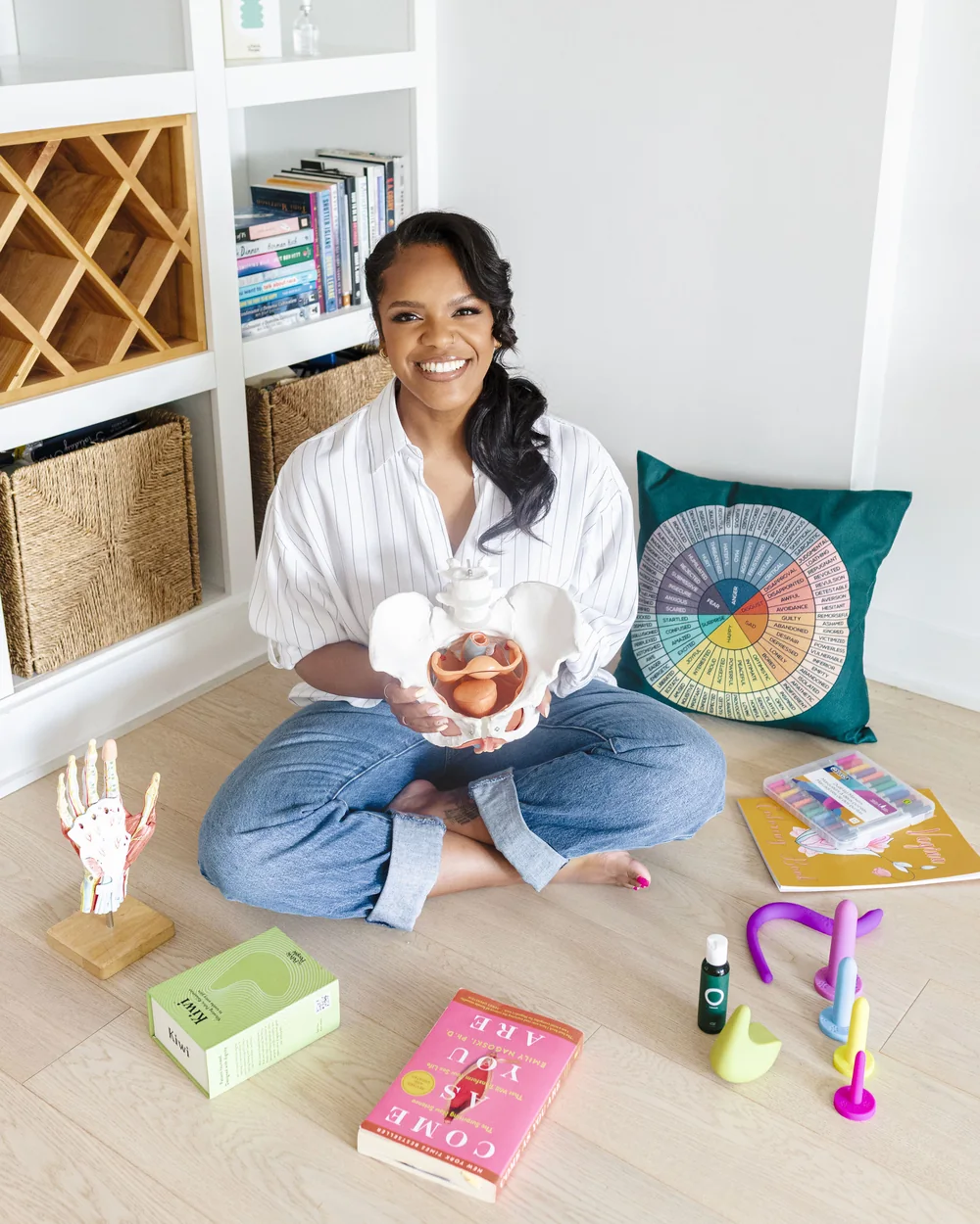 Karina Jackson, assise au sol, souriante, son modèle anatomique de bassin dans les mains, entourée de livres, d'un coussin à roue des émotions et d'outils de santé pelvienne