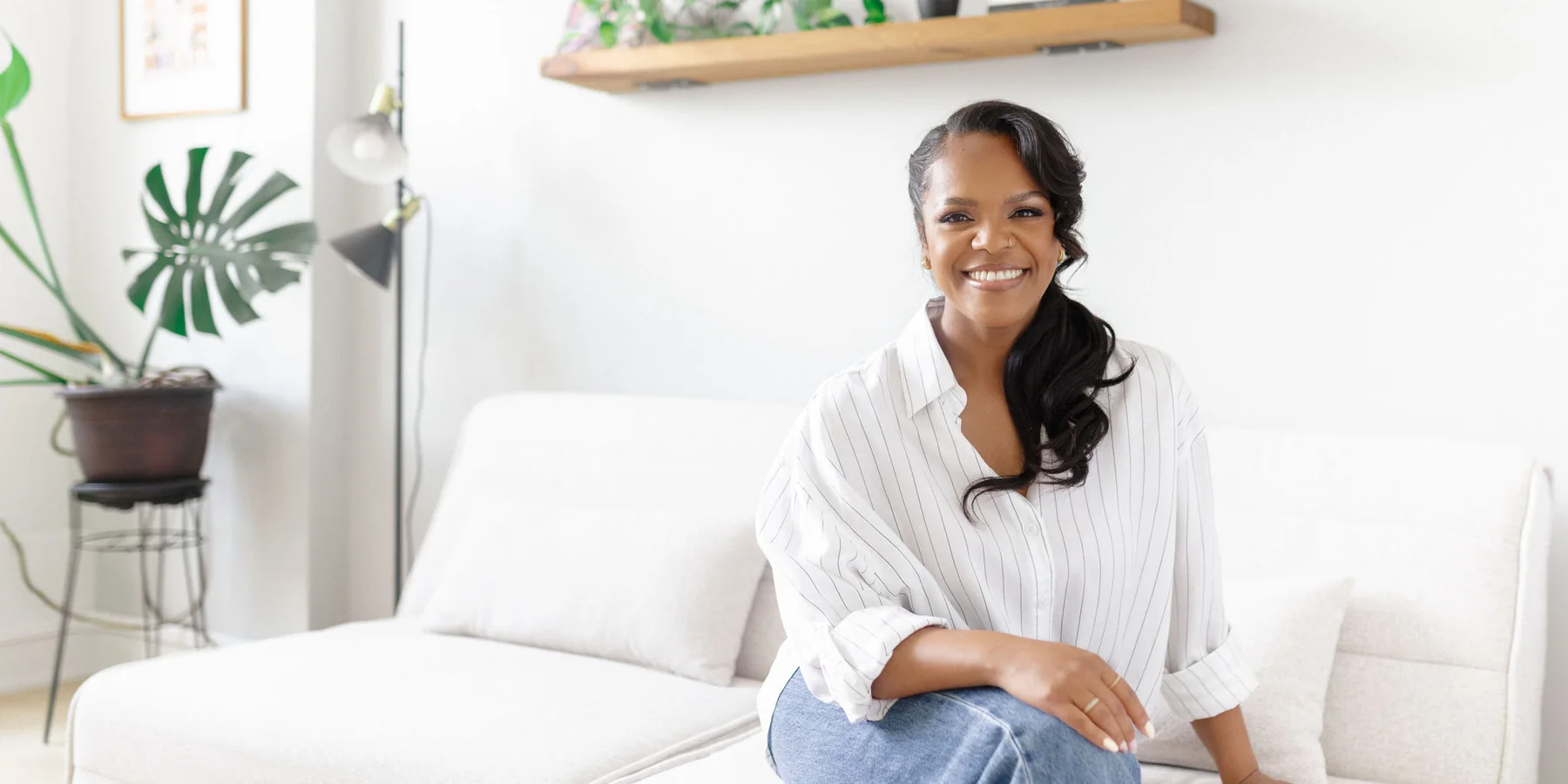 Karina Jackson seated on a white sofa in a bright room, smiling, with wooden shelves, plants and a large monstera