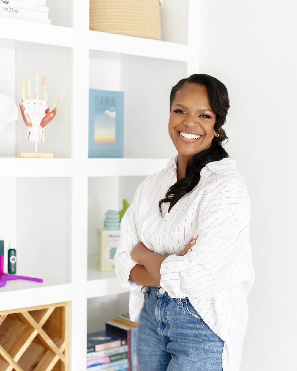 Karina Jackson, debout et souriante, les bras croisés devant sa bibliothèque blanche, où repose son modèle anatomique de main